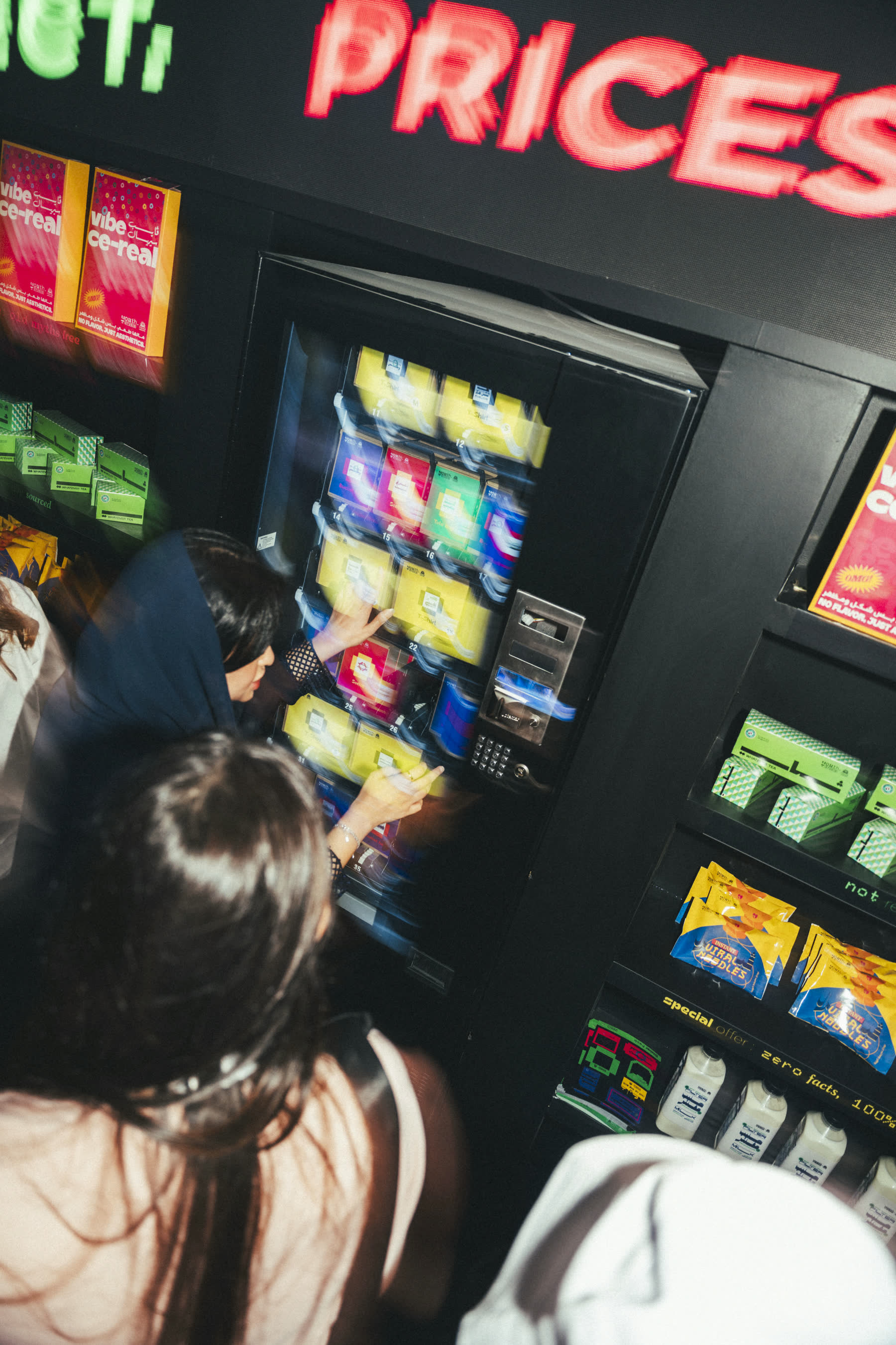 Visitors at the myth vending machine under a neon PRICES sign
